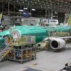 An interior view of a large aircraft assembly plant showcasing multiple Boeing 737 Max airplanes under construction. The green protective coating covers the aircraft fuselages, and various assembly platforms and scaffolding surround the planes. Workers in safety gear are seen inspecting and working on the airplanes in the brightly lit facility.