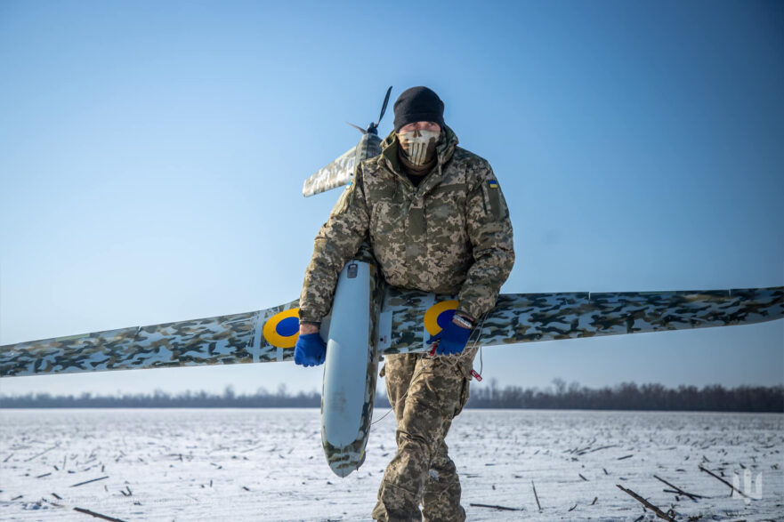 An operator carries a Ukrspecsystems UAV over snow-covered ground against a blue sky.