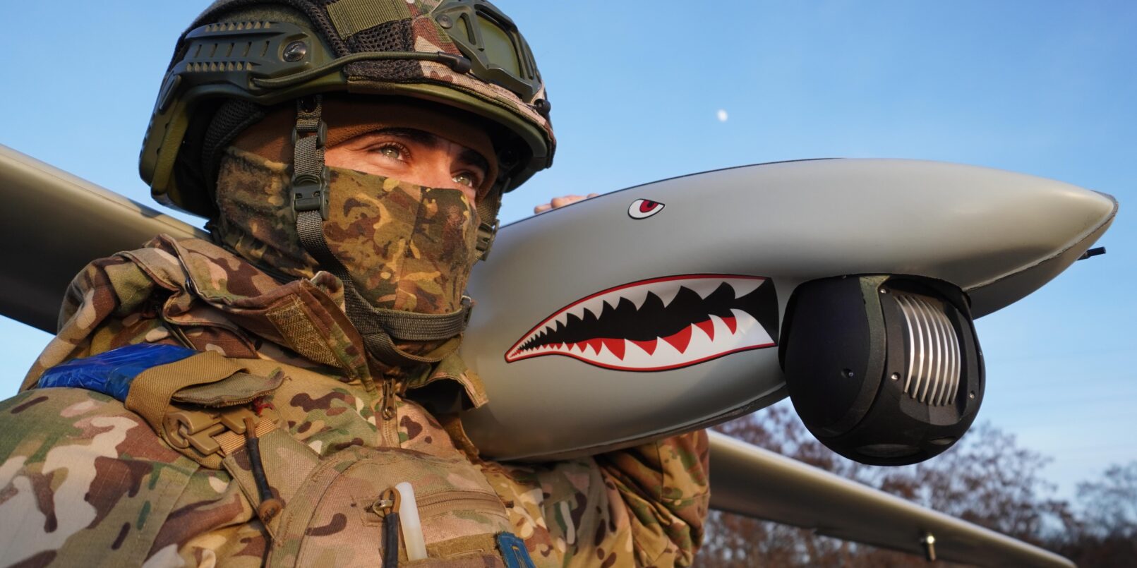 A Ukrainian soldier holds a Ukrspecsystems Shark UAV over his shoulder against a blue sky