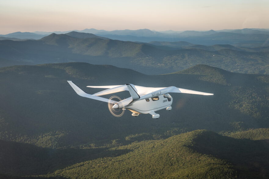 N916LF, a white electric airplane made by Beta Technologies, shown in flight over green hills with a full disc capture of its spinning rear propeller