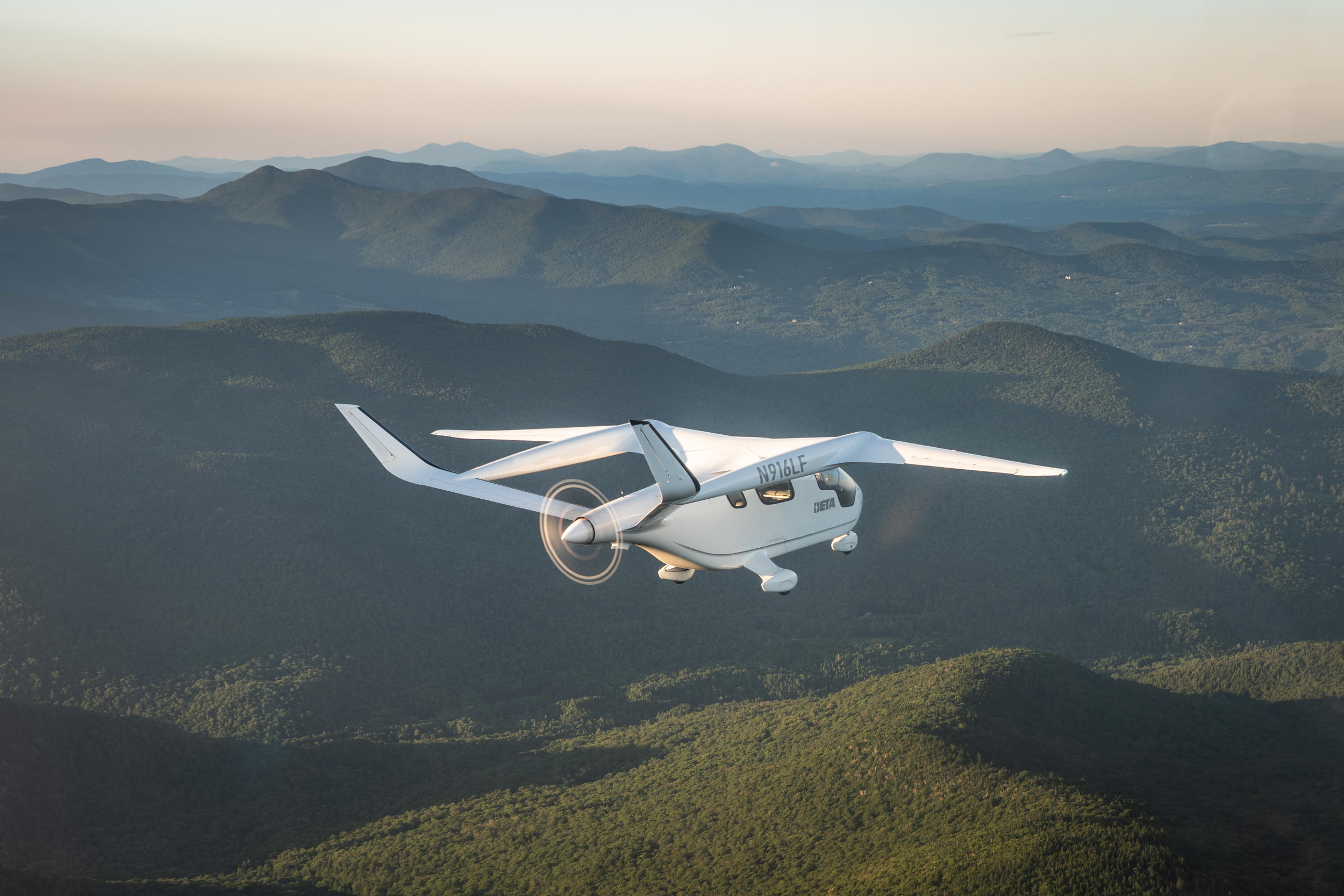 N916LF, a white electric airplane made by Beta Technologies, shown in flight over green hills with a full disc capture of its spinning rear propeller